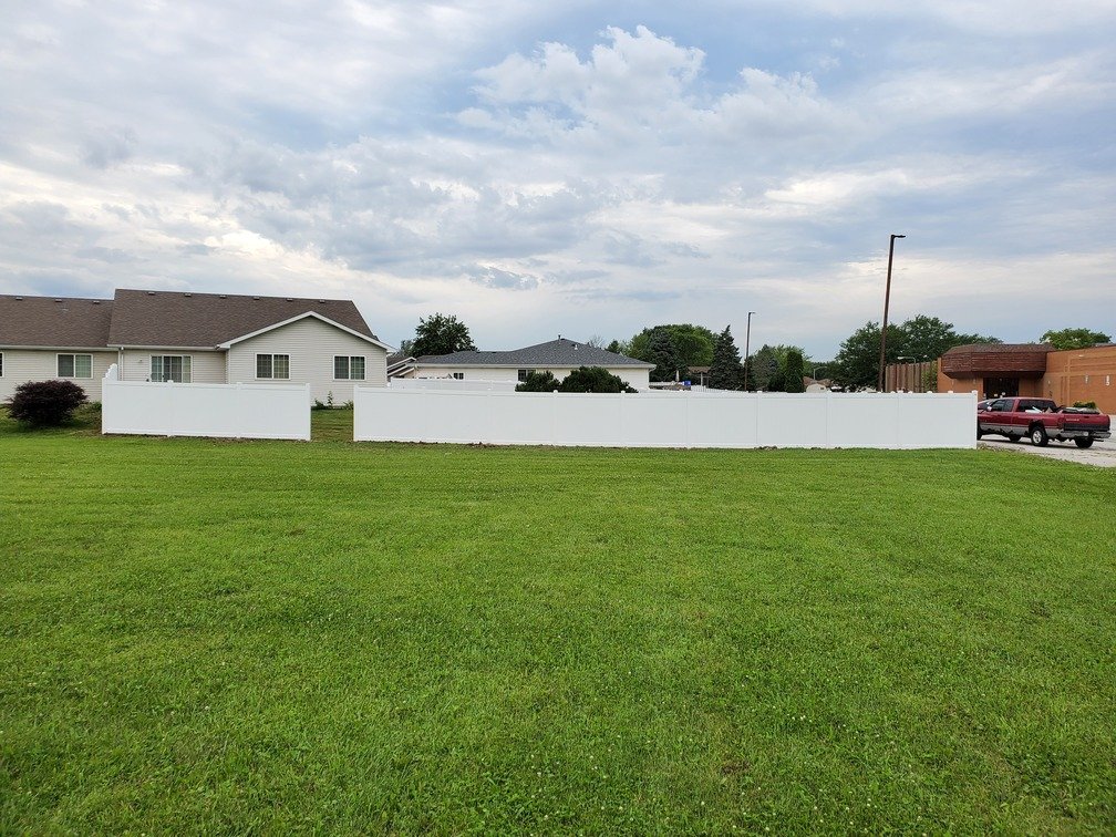 A grassy field with a white fence extending across the background, residential houses and cars in the distance, under a cloudy sky.