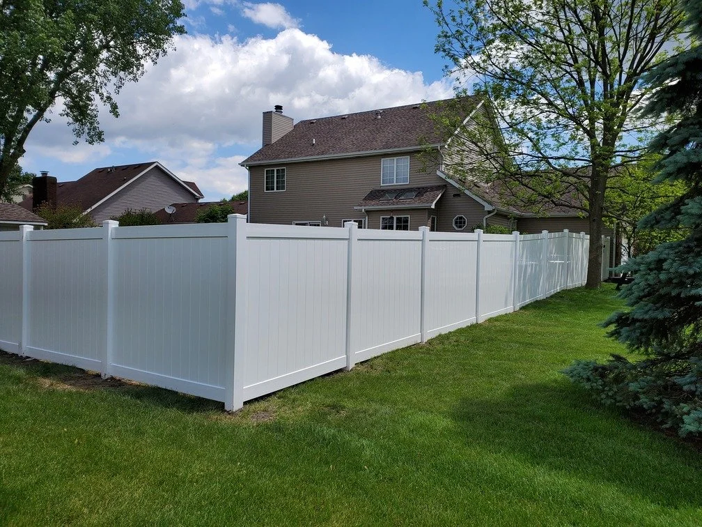 A white privacy fence surrounds a backyard with green grass, a large tree, and neighboring houses under a partly cloudy sky.