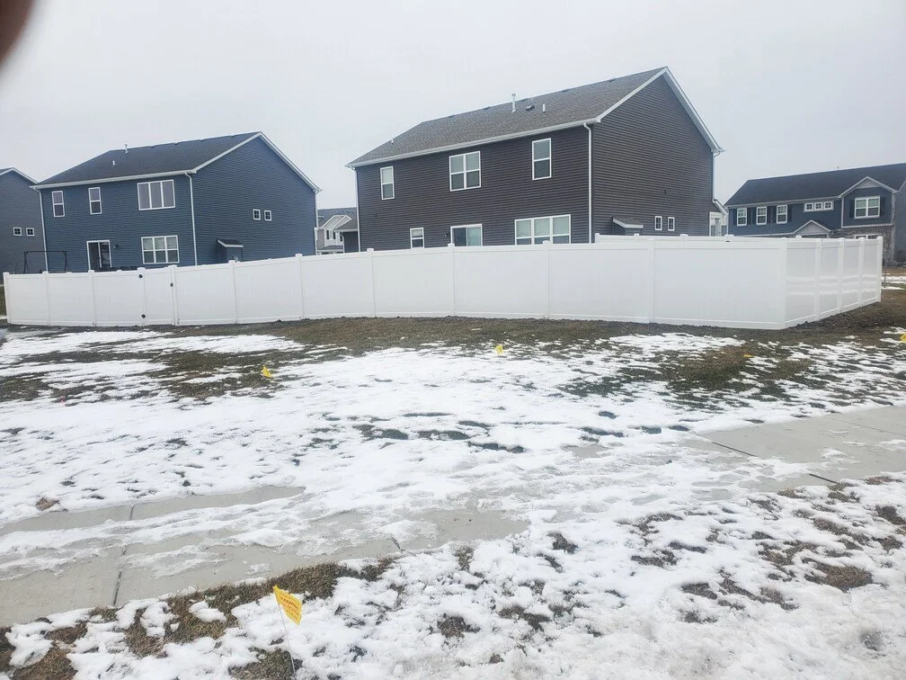 A fenced backyard with snow patches on the ground, surrounded by several modern houses with dark siding and large windows.