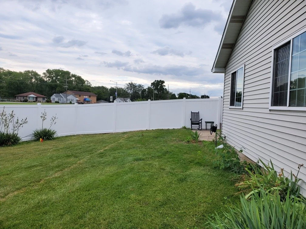 Backyard with green grass, a white vinyl fence, a patio with two chairs, a house with beige siding, and neighboring houses in the distance under a cloudy sky.