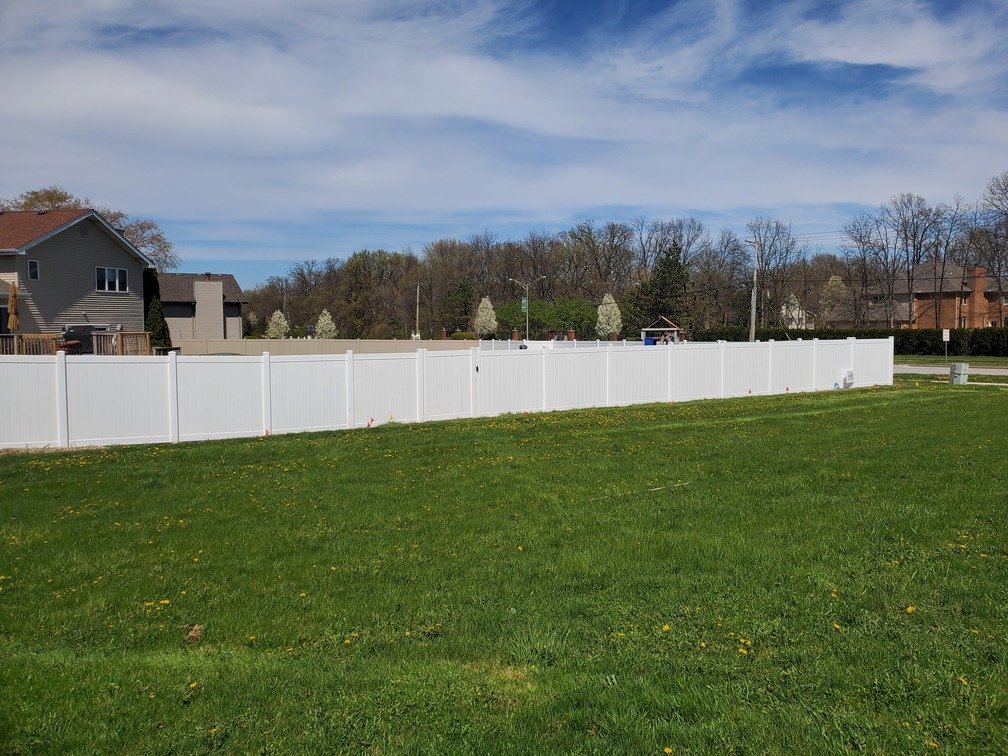 A white fence separating a grassy yard from neighboring houses and trees under a partly cloudy blue sky.