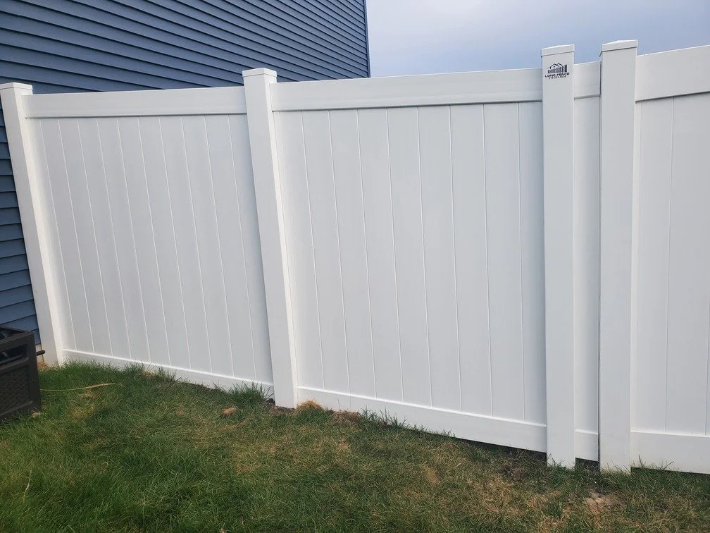 White privacy fence with vertical panels and support posts adjacent to a blue house with vinyl siding, on a grassy yard.