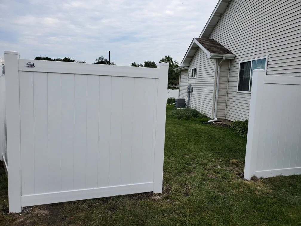 View of a white vinyl privacy fence next to a house with beige siding, a small window, and an air conditioning unit in the yard.