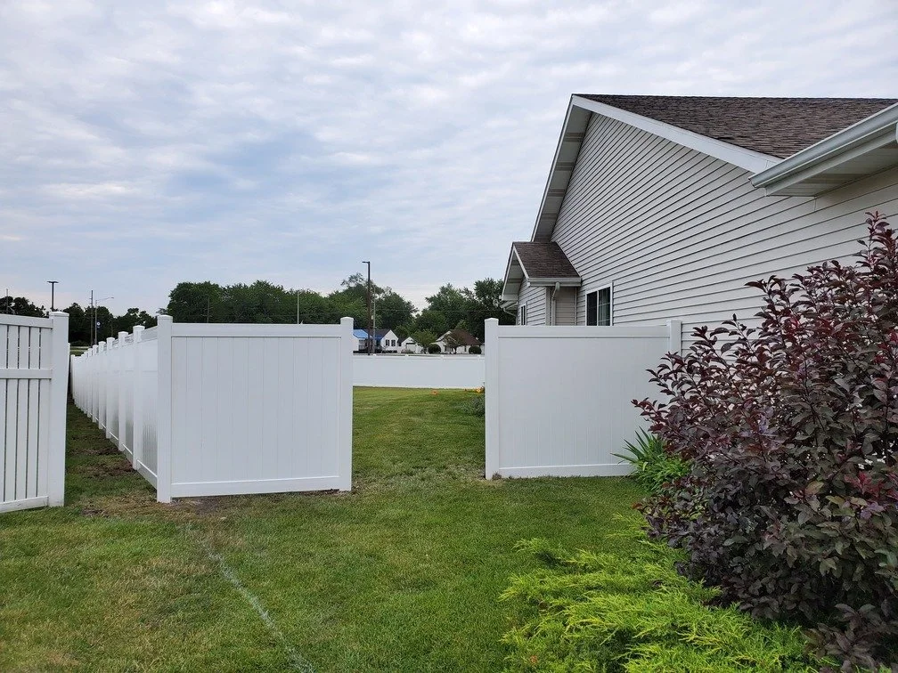 A backyard with a white vinyl fence, a house with beige siding, green grass, and some bushes with reddish leaves.