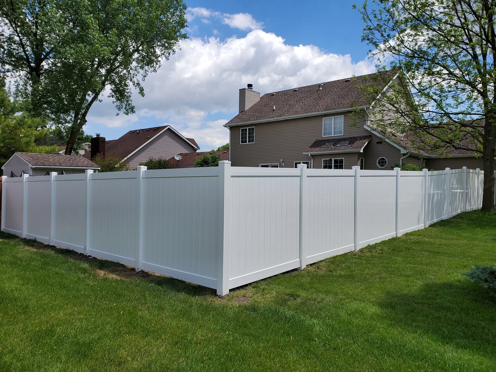 A backyard with a white privacy fence, green grass, trees, and a two-story house with a gray roof and beige siding under a partly cloudy sky.