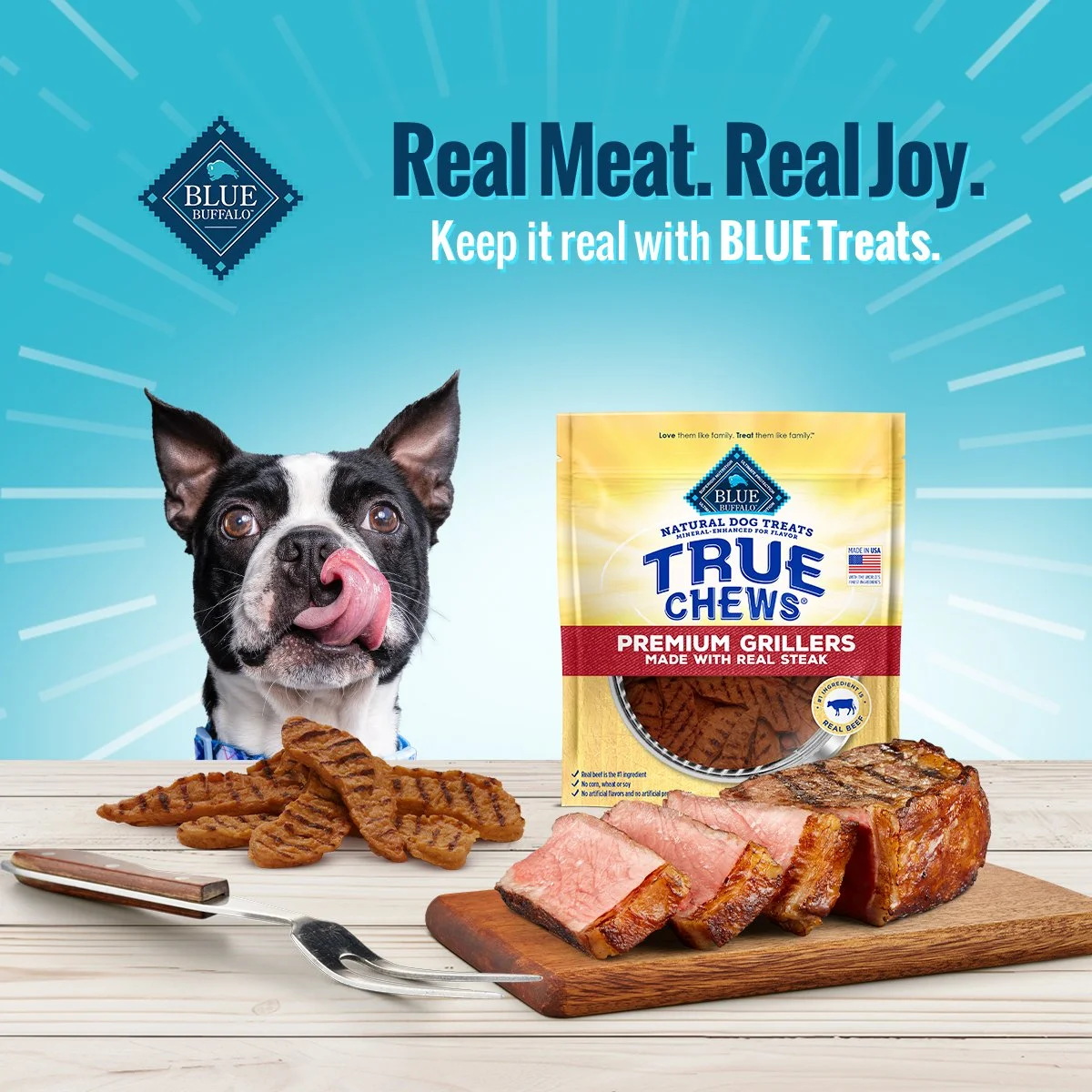 A boston terrier licks his lips with eyes wide. In front of him is a plate of grilled treats. To his right is a bag of BLUE True Chews and a cutting board with sliced steak on top.