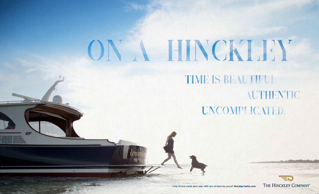 A woman walks off the back of a Hinckley yacht onto a sandbar where her labrador is jumping up to greet her. Their silhouettes are dark against a bright blue sky and the line says "On a Hinckley, time is beautiful. Authentic. Uncomplicated."