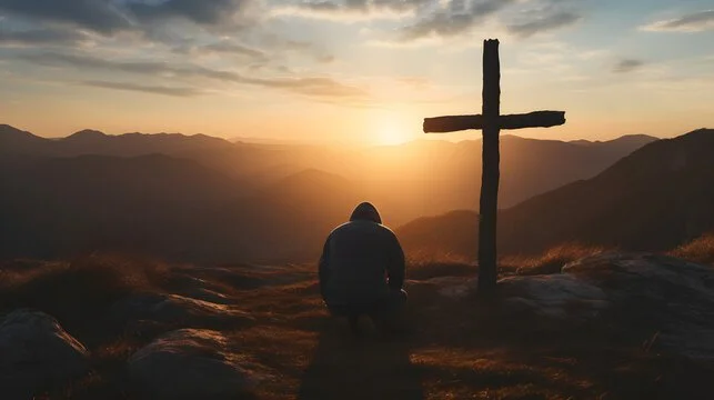Person praying or reflecting next to a wooden cross during sunset on a mountain landscape.