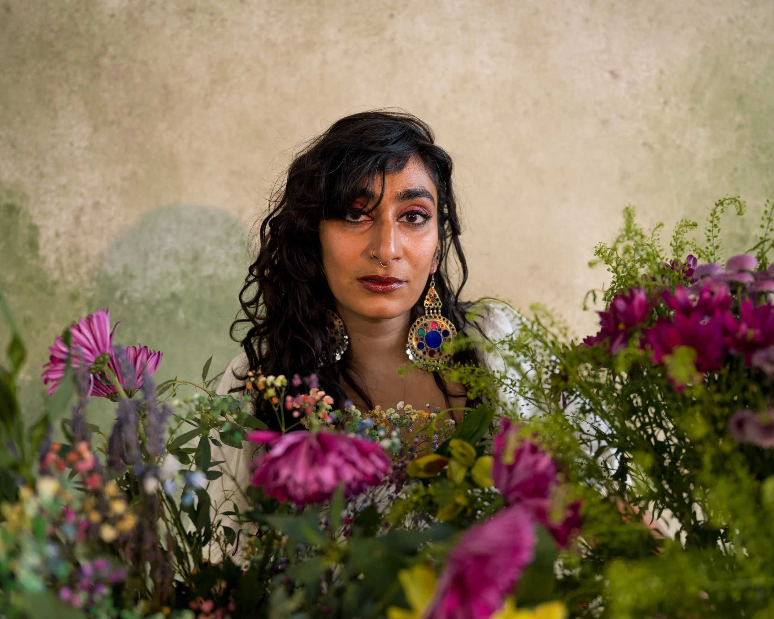 Fatimah Asghar with dark curly hair wearing colorful earrings, surrounded by pink and purple flowers, with a beige textured background.