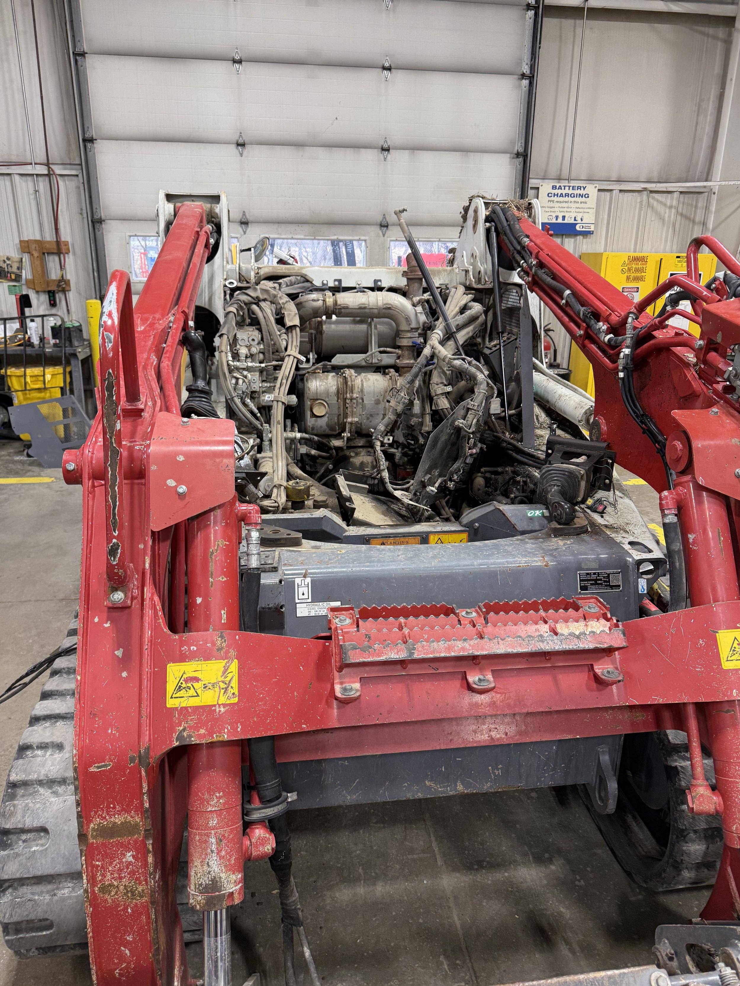 A construction vehicle without an enclosed cab, showing an exposed engine and hydraulic system, inside a workshop or garage with warning signage and equipment on the wall.