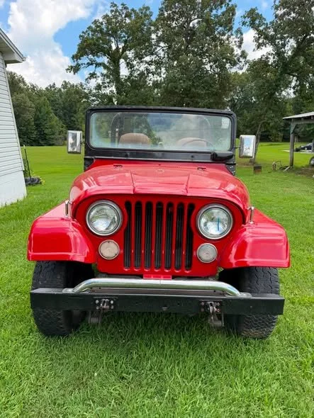 Red vintage Jeep with black front grille parked on green grass with trees and a house in the background.