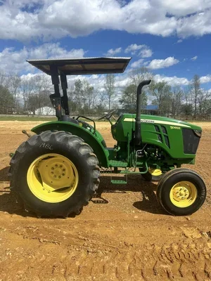 Green tractor with a canopy on a dirt field, with trees and a partly cloudy sky in the background.