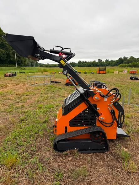 Orange and black compact tracked loader with a lifting arm and a bucket attachment, positioned on grassy terrain in an open field.
