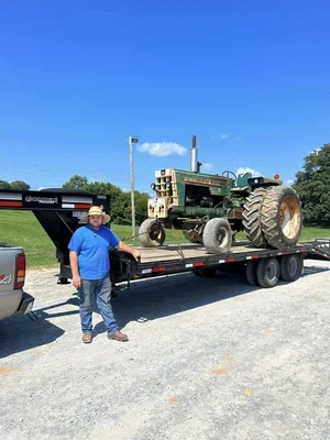 Man standing next to a flatbed trailer carrying an old tractor on a gravel lot under a blue sky.