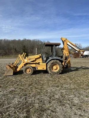 A yellow backhoe loader parked on grass, with a clear blue sky and trees in the background.