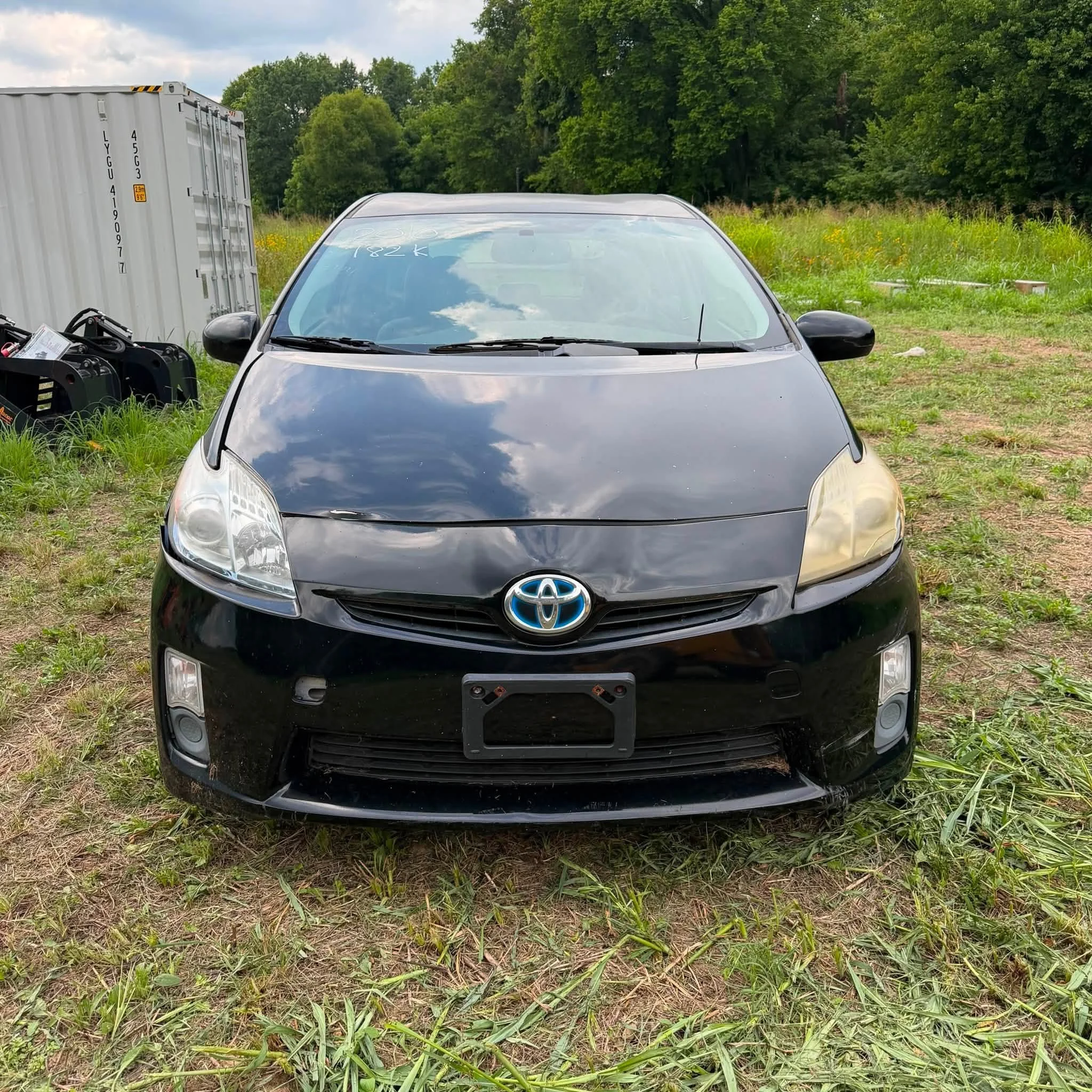 A black Toyota car parked on grassy dirt area with a large container on the left and green trees in the background.