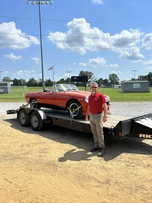 A man in a red shirt standing next to a red vintage car on a flatbed trailer at a sports field with clouds and blue sky in the background.