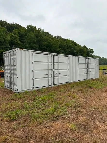 A gray shipping container lying on grassy ground in an open field with trees and cloudy sky in the background.