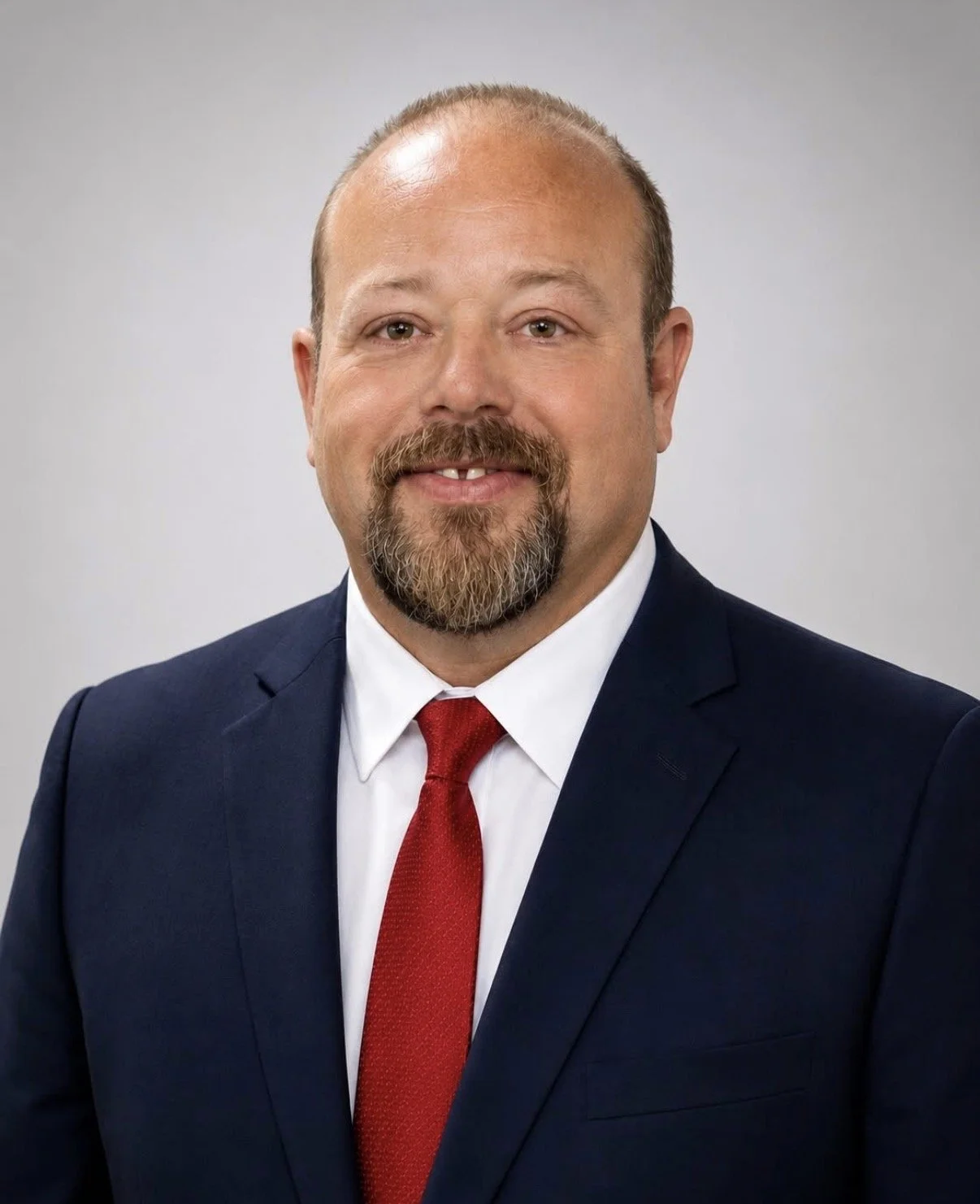 A middle-aged man with a beard and mustache, wearing a dark blue suit, white shirt, and red tie, posing for a professional headshot against a light gray background.