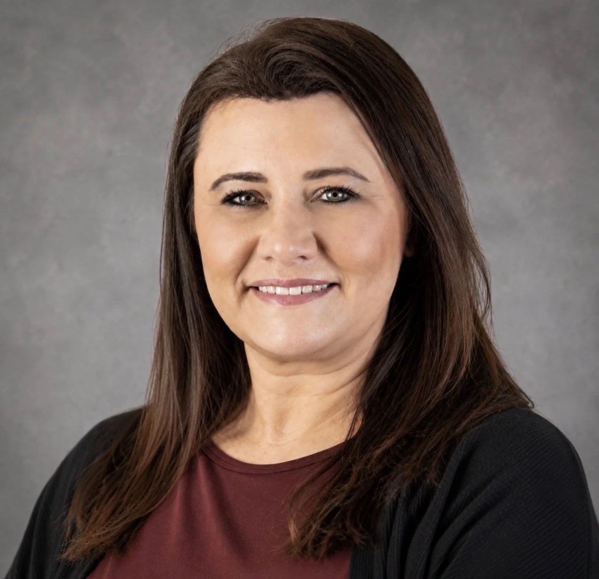 Professional headshot of a woman with shoulder-length brown hair, wearing a maroon top and black blazer, smiling against a gray background.