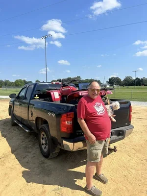 Man in sunglasses and red T-shirt standing beside a black pickup truck on a dirt field, holding a small dog, with a sports field and blue sky in the background.