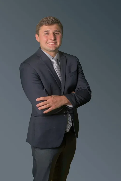 A young man in a navy blue suit with a light tie, smiling with arms crossed, standing against a plain gray background.