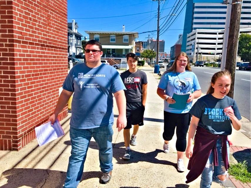 Four young people walking on a city sidewalk on a sunny day, carrying notebooks and wearing casual clothes.