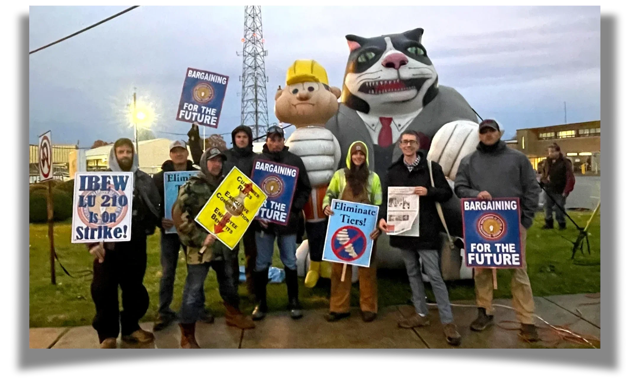 A group of people stand with signs which read "IBEW LU 210 Is on Strike" in front of a large inflatable cat