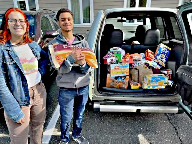 A man and woman smile next to a trunk filled with food