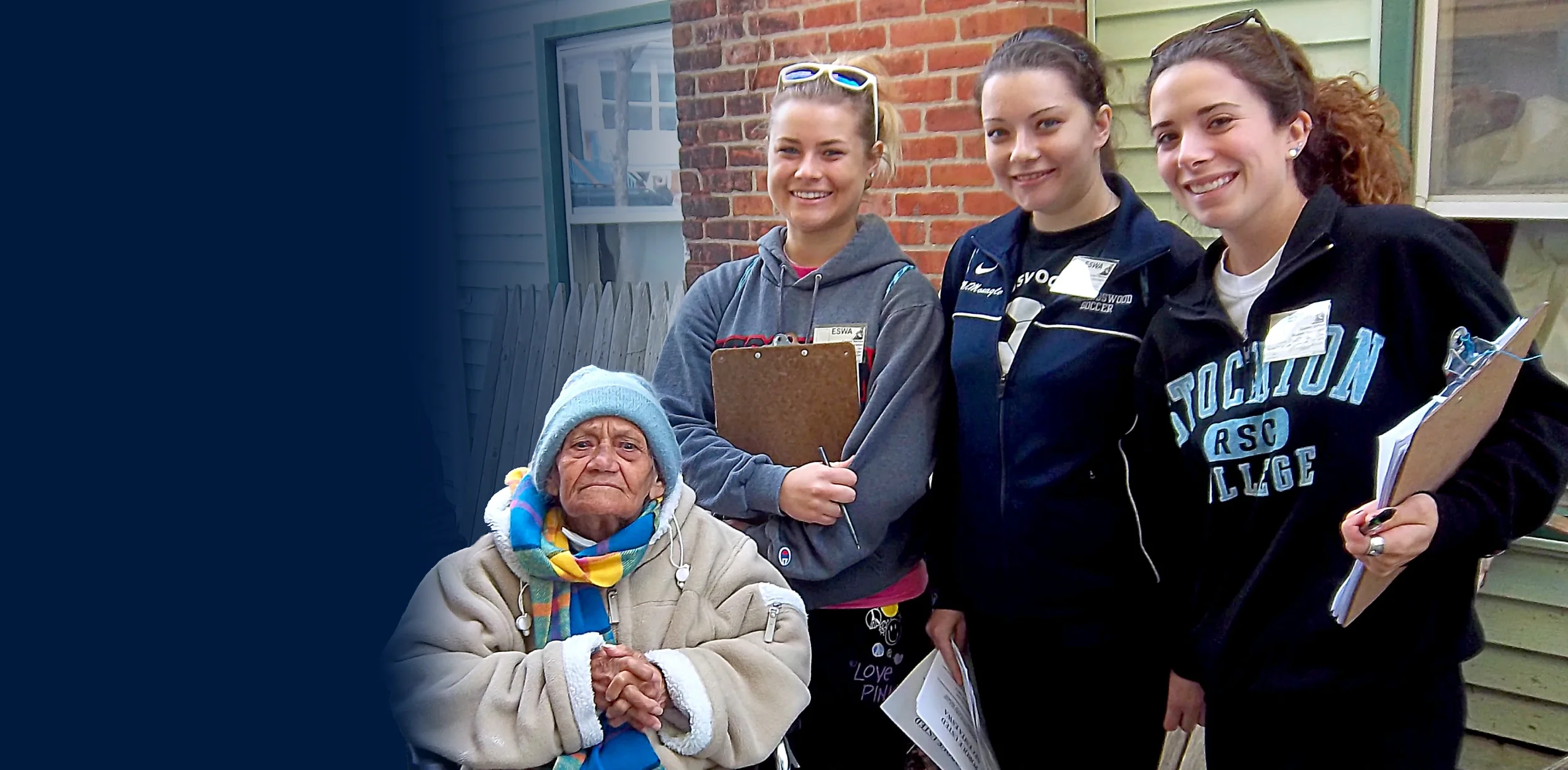 Four young women and one elderly woman standing together outside, smiling, in front of a brick and green house, with the elderly woman seated and wearing a winter hat and scarf.