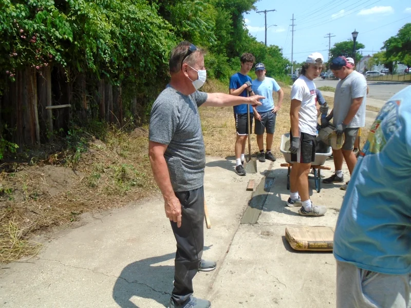 A man directs a group of people who have shovels and a wheelbarrow