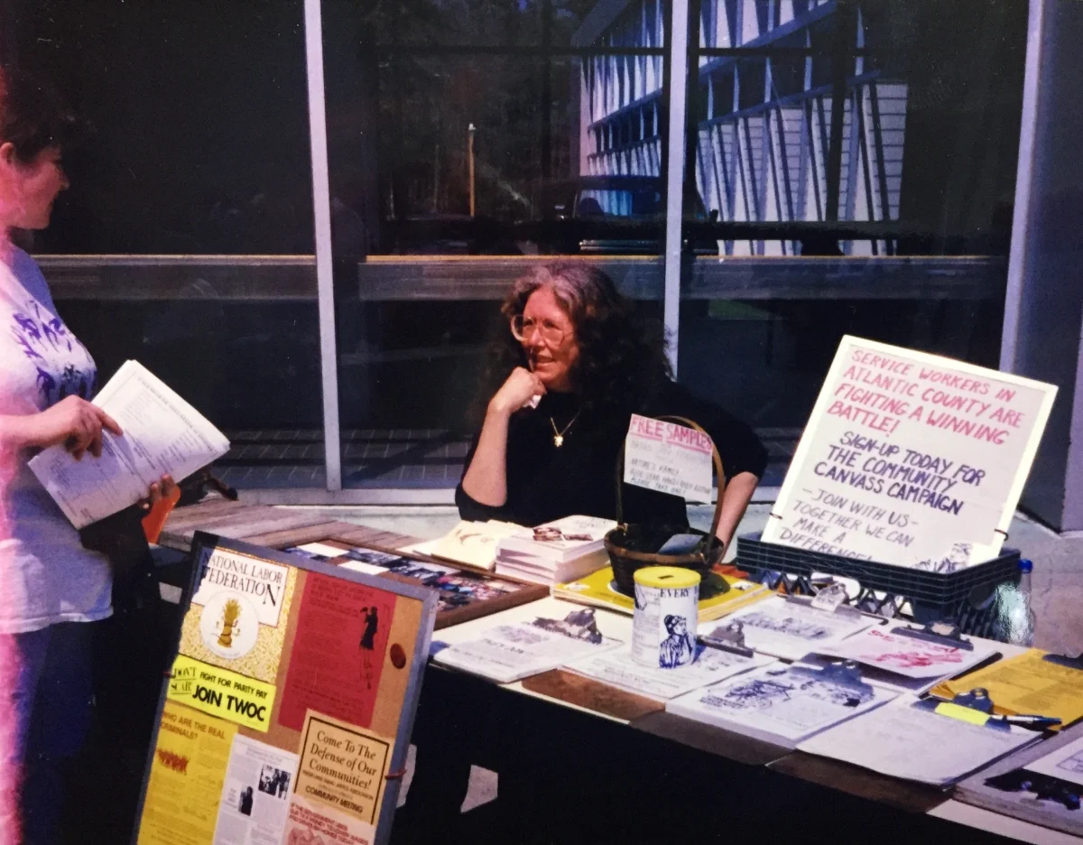 A woman sitting down next to a table with a sign that reads "Service Workers are fighting a winning battle!"
