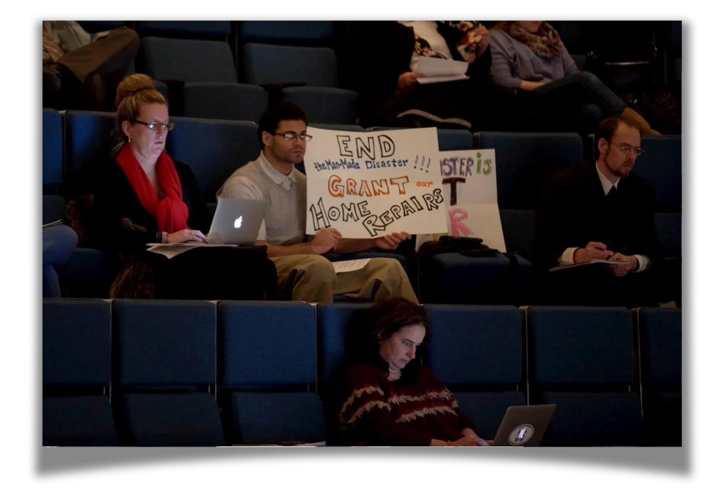 A man holds a sign which reads "End The Man-Made Disaster!!! Grant our Home Repairs"