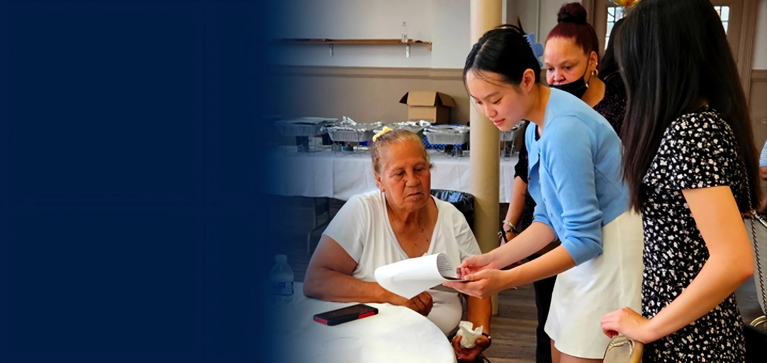 A group of people gathered around an elderly woman sitting at a table, reviewing a document.