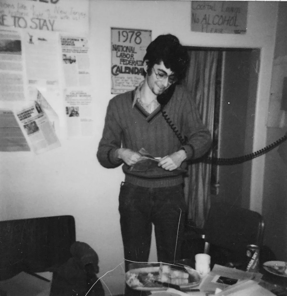 A black and white photo of a young man on the phone with a poster that reads "1978"