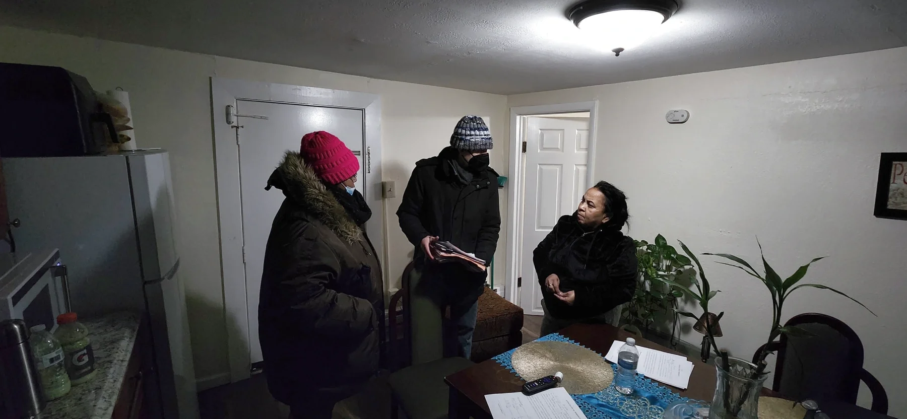 Three people talk to one another inside while wearing heavy winter coats and hats