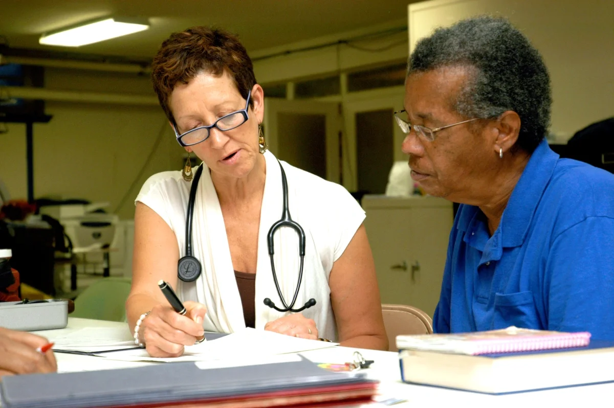 A female doctor with a stethoscope around her neck is writing on a document, while a male patient in a blue shirt sits next to her, looking at the document.