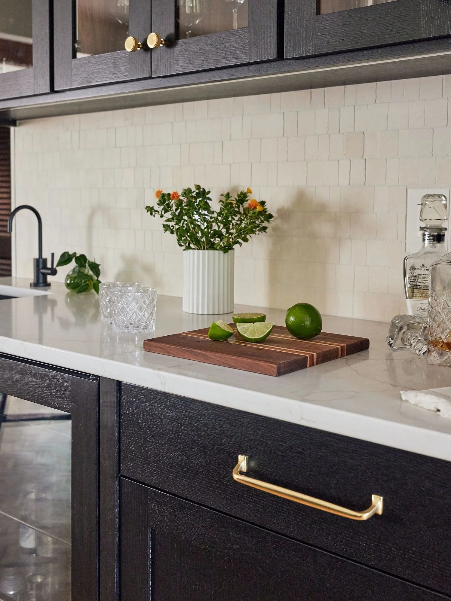 Black stained oak cabinet with brass handles, white marble countertop, rustic white subway tile backsplash, potted plant, three lime slices and whole lime on cutting board, glass decanter, and glass cup on the counter.
