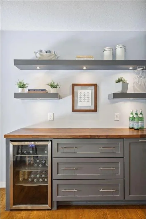 Kitchen bar area with gray cabinets, a wooden countertop, floating shelves with plants, books, and decorative items, and a mini wine fridge.