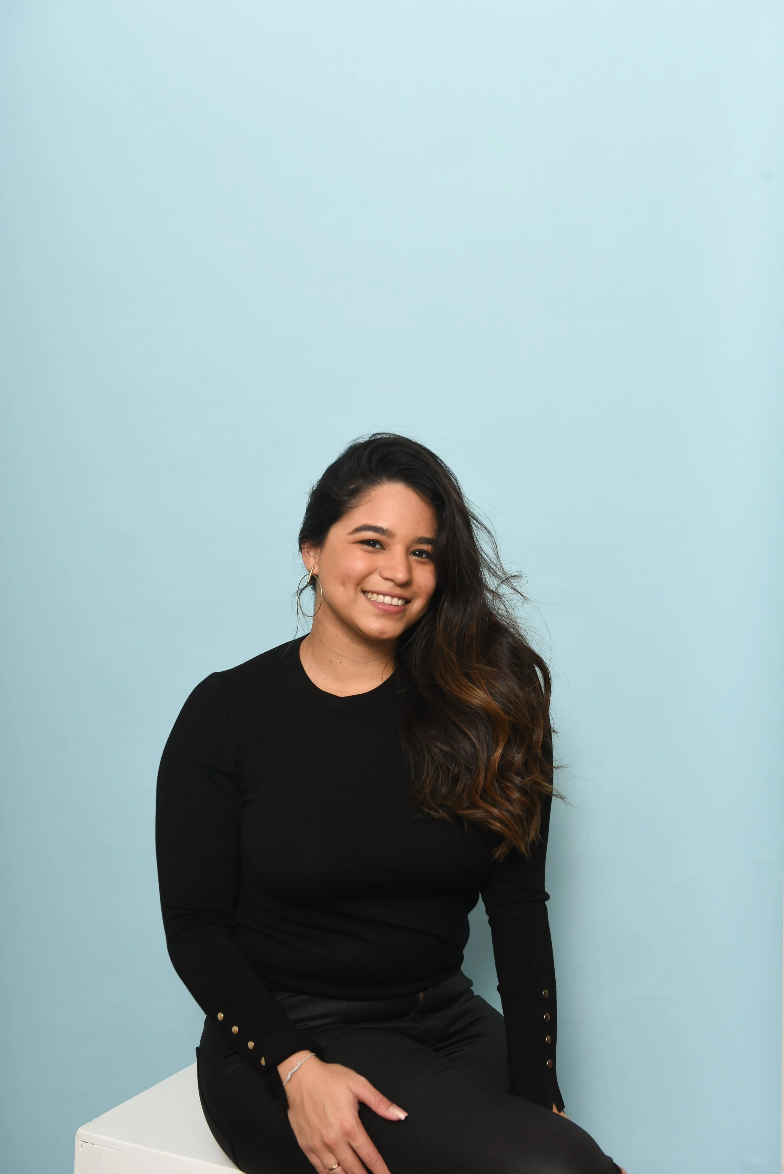 A woman with long dark hair and light skin, smiling and sitting on a white surface against a plain light blue background, wearing a black long-sleeve top and black pants.