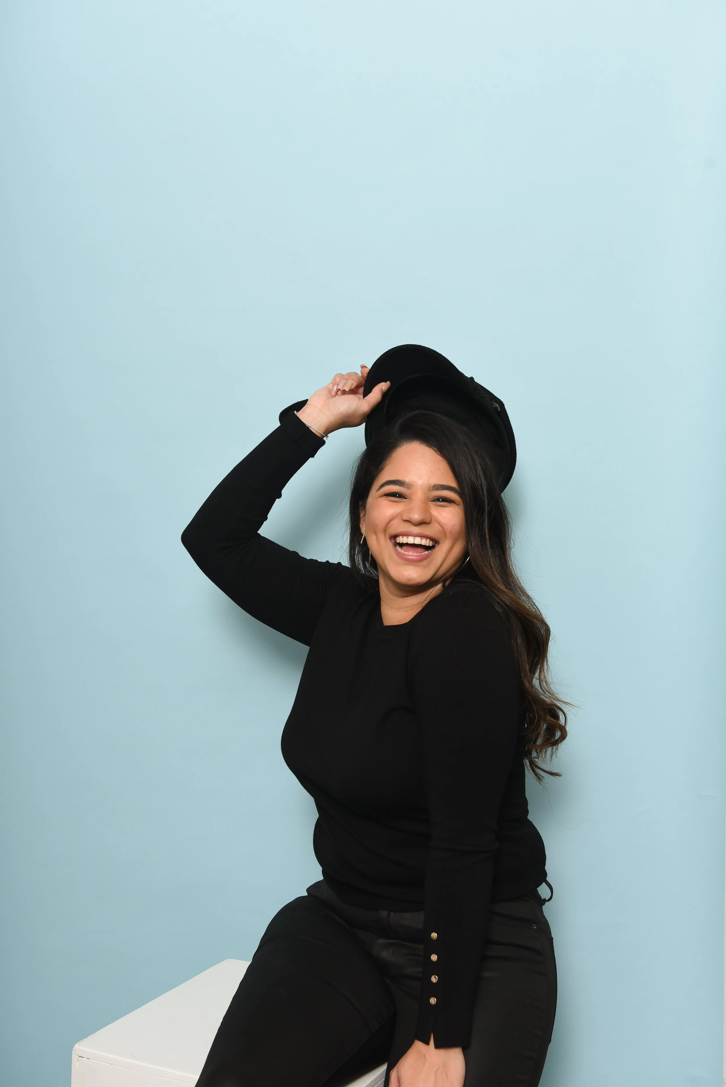 A woman with long dark hair sitting on a white block against a light blue wall, holding a black cap, smiling.