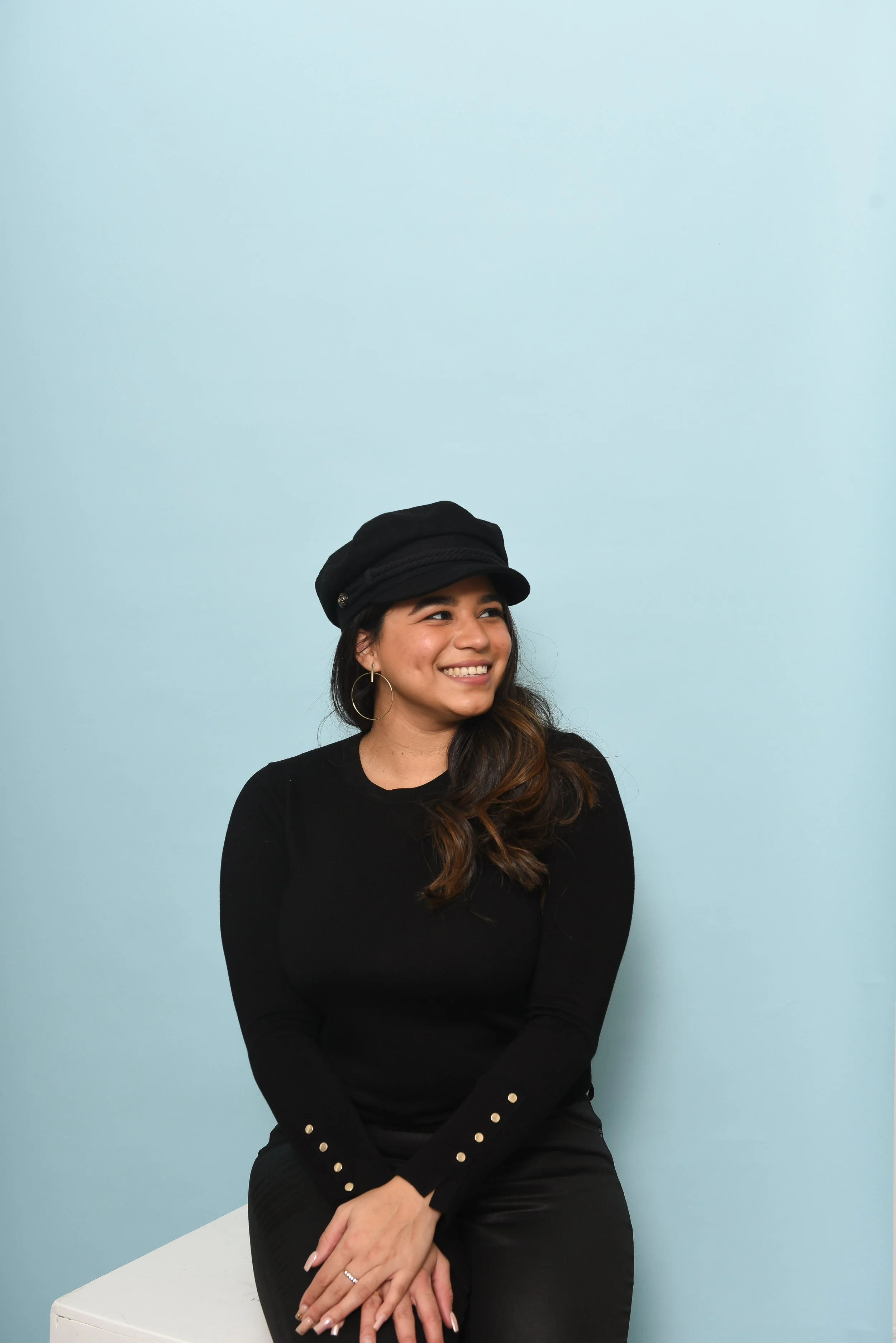 A woman sitting on a white box against a light blue wall, wearing a black hat and a black long sleeve shirt with gold buttons on the sleeves, looking away and smiling.