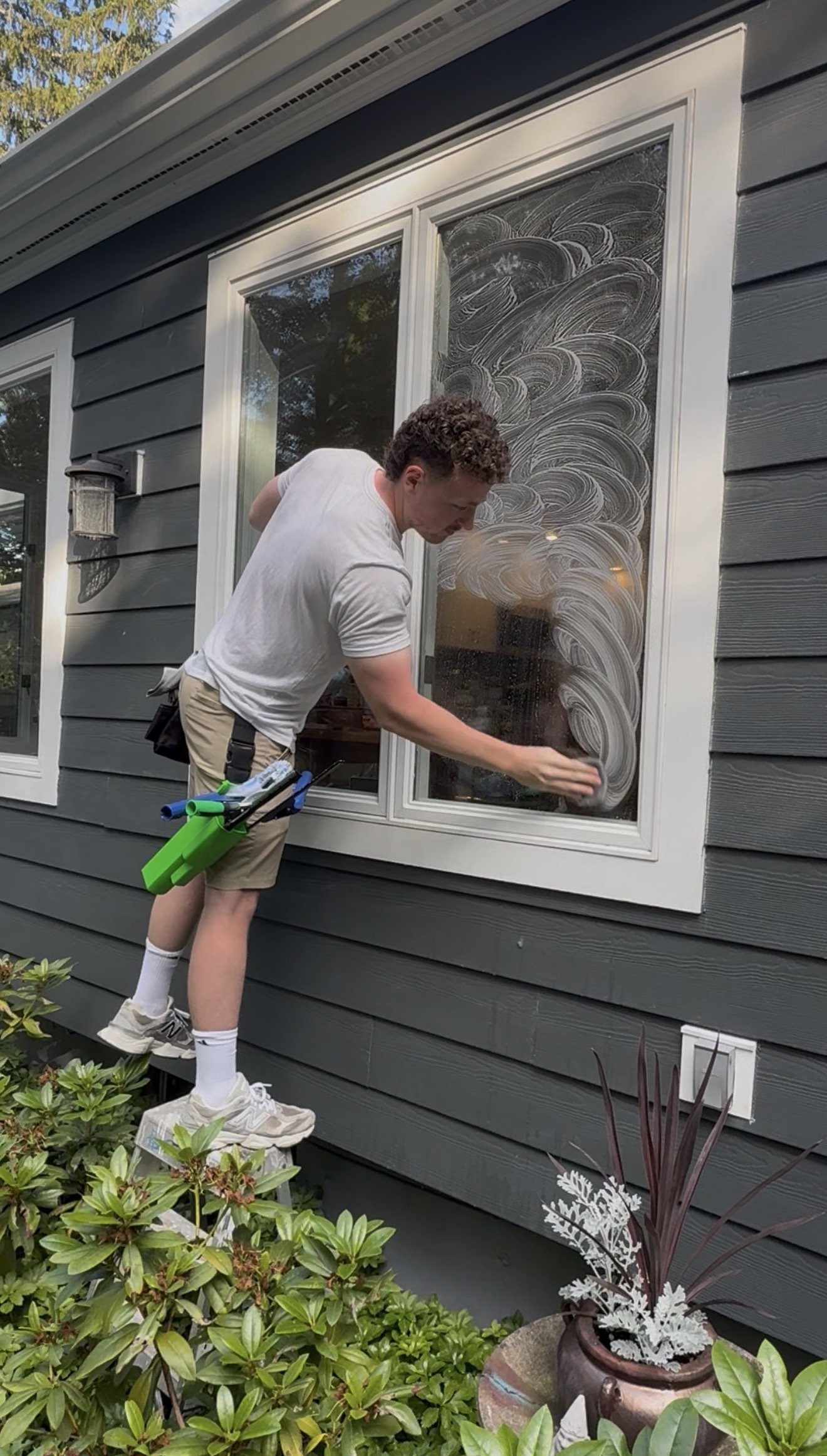 A young man cleaning the exterior window of a house with soap and water, standing on a stool outside a dark gray house with white trim, surrounded by green plants.