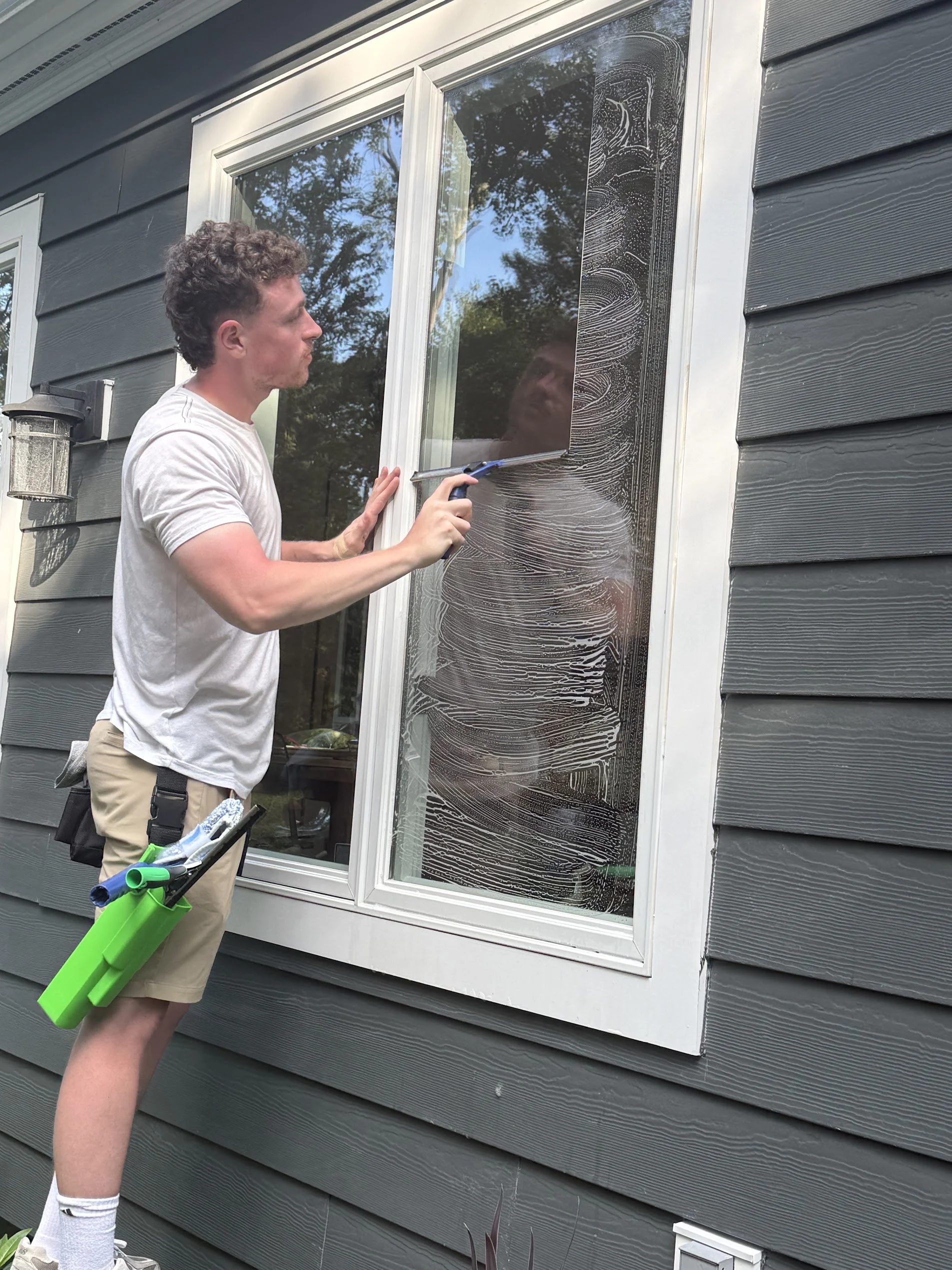 A man cleaning a window with a squeegee from inside the house, facing the glass, with cleaning foam on the window. The house has gray horizontal siding, and the man is wearing a white t-shirt, beige shorts, white socks, and shoes, with cleaning tools hanging from his pocket.