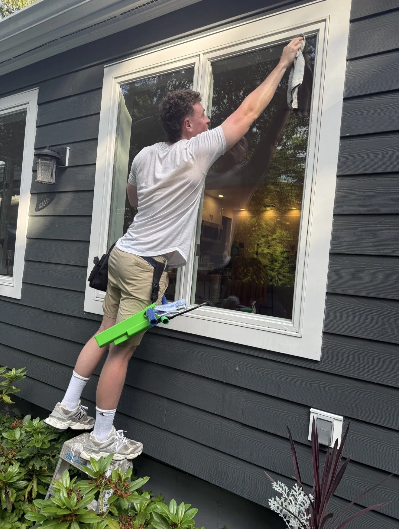 A man standing on a small ladder cleaning a window on the exterior of a house with dark gray siding.