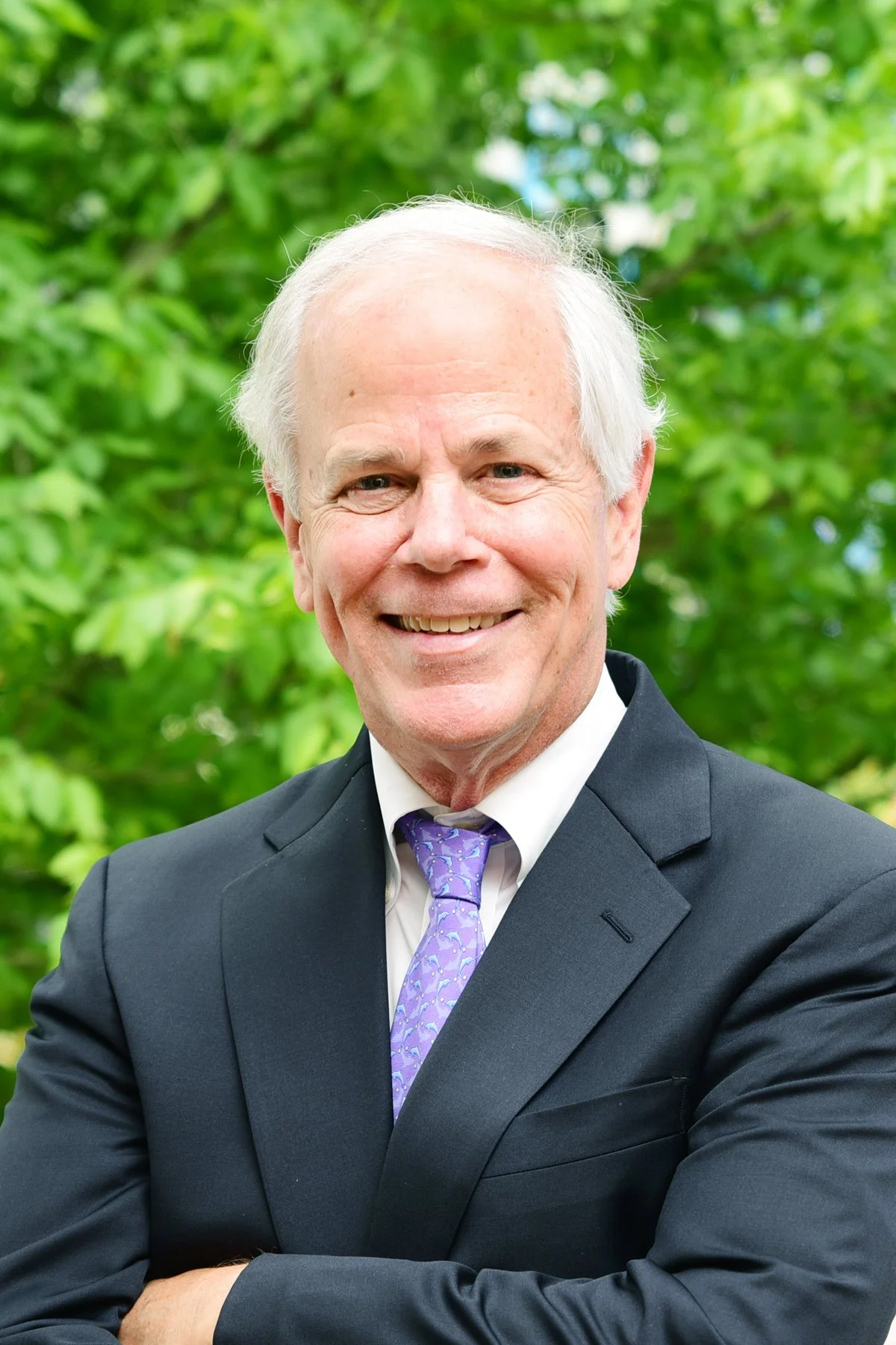An elderly man in a dark suit with a purple patterned tie smiling outdoors with green foliage in the background.