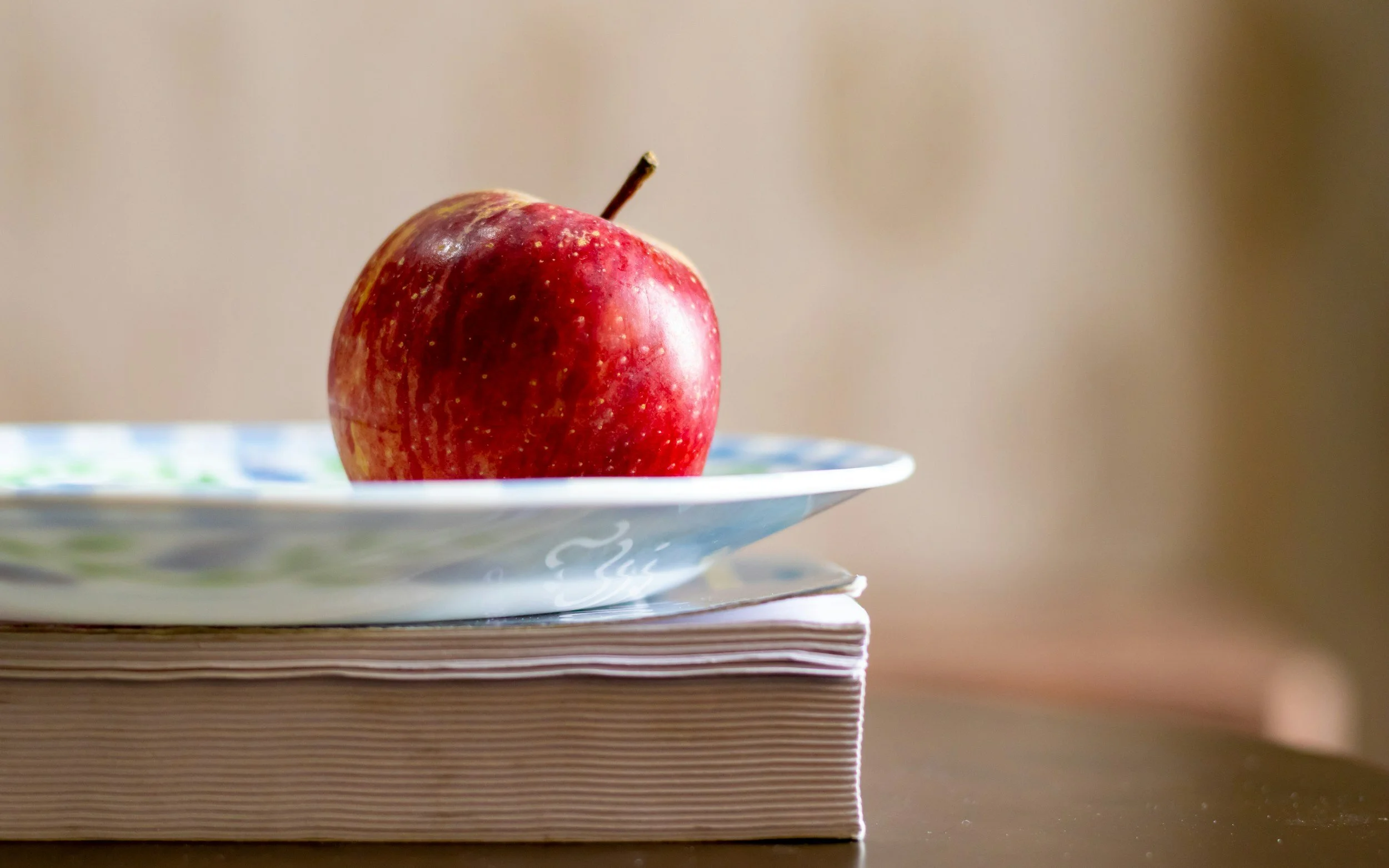 Close-up of a red apple with some yellow spots, placed on a decorative plate, resting on a stack of papers or books.