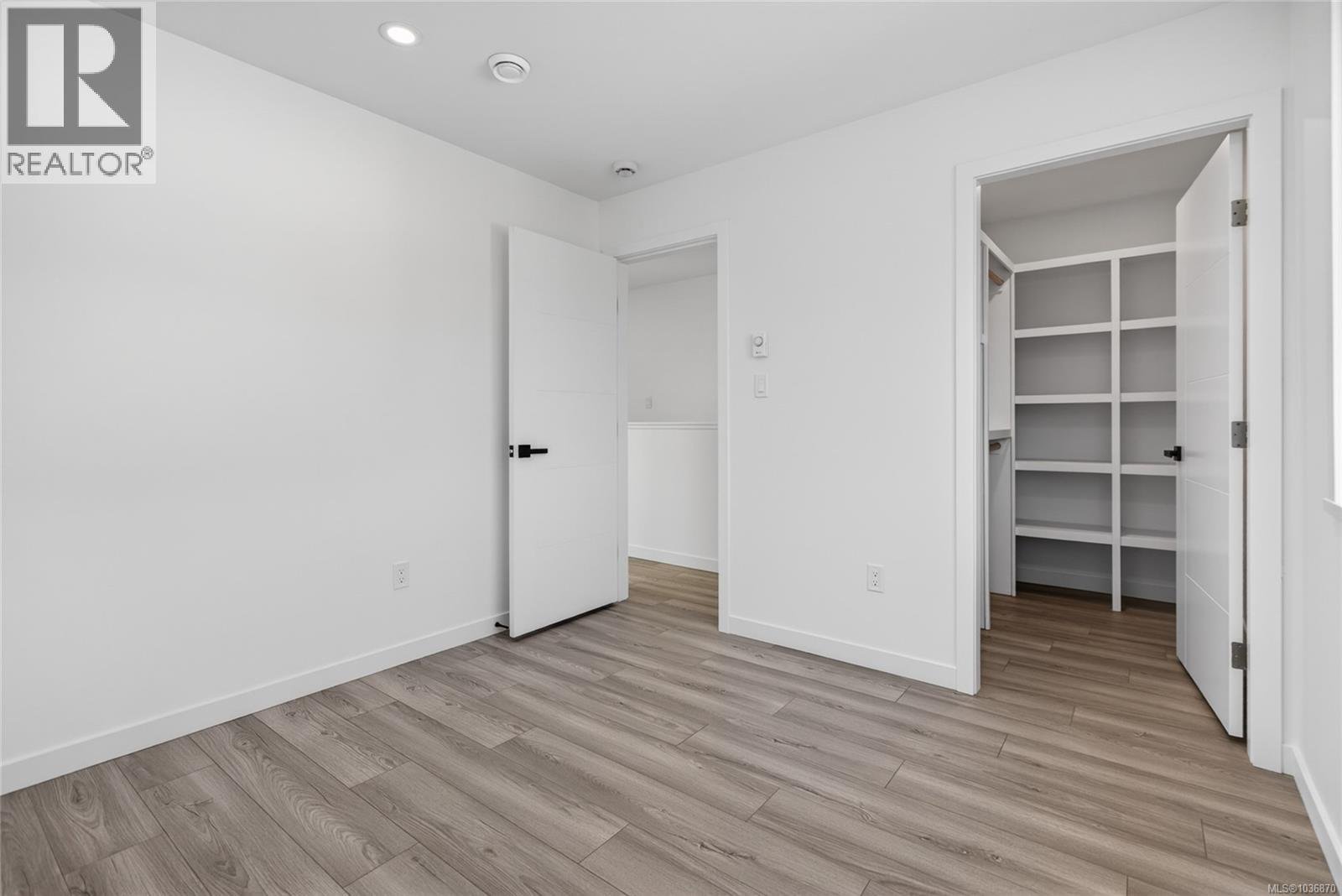 Empty room with white walls, wooden floor, open closet with shelves, and a door leading to another space.