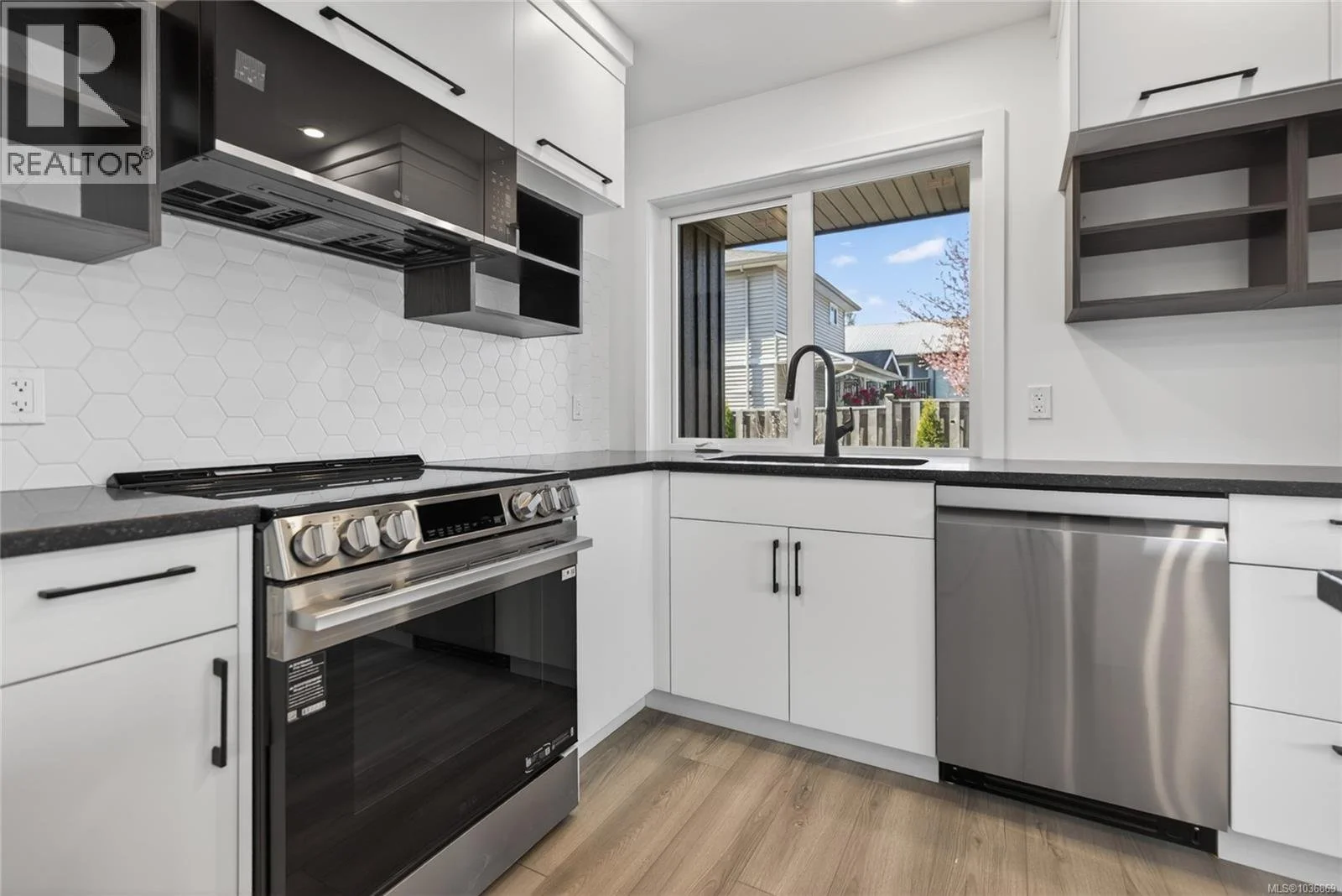 Modern kitchen with white cabinets, black countertops, stainless steel appliances, a window overlooking a backyard, and open shelving.