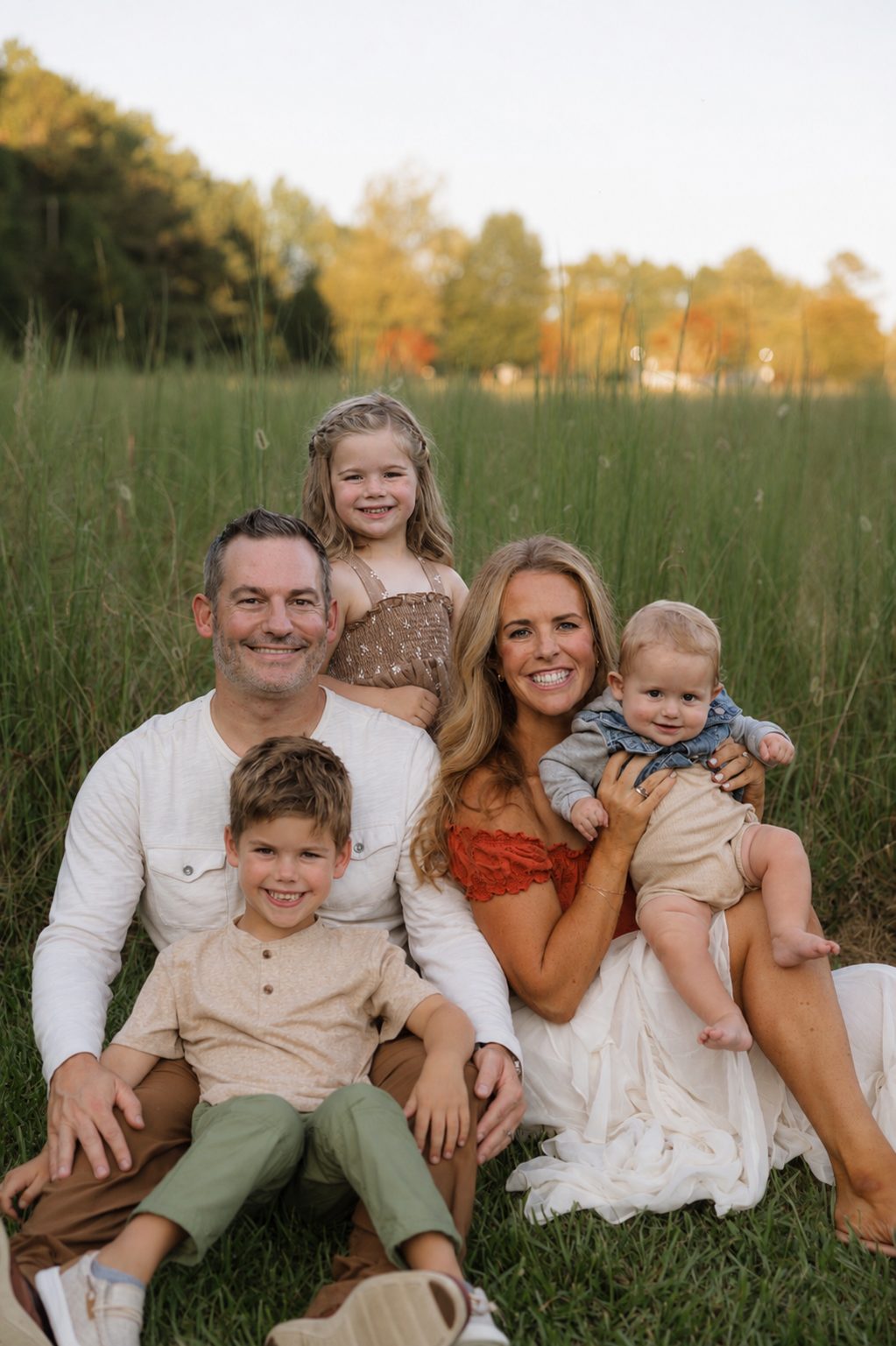 A smiling family of five sitting on grass in a field during golden hour, with trees in the background. The family includes man and woman with three children, two boys and one girl, all dressed casually.